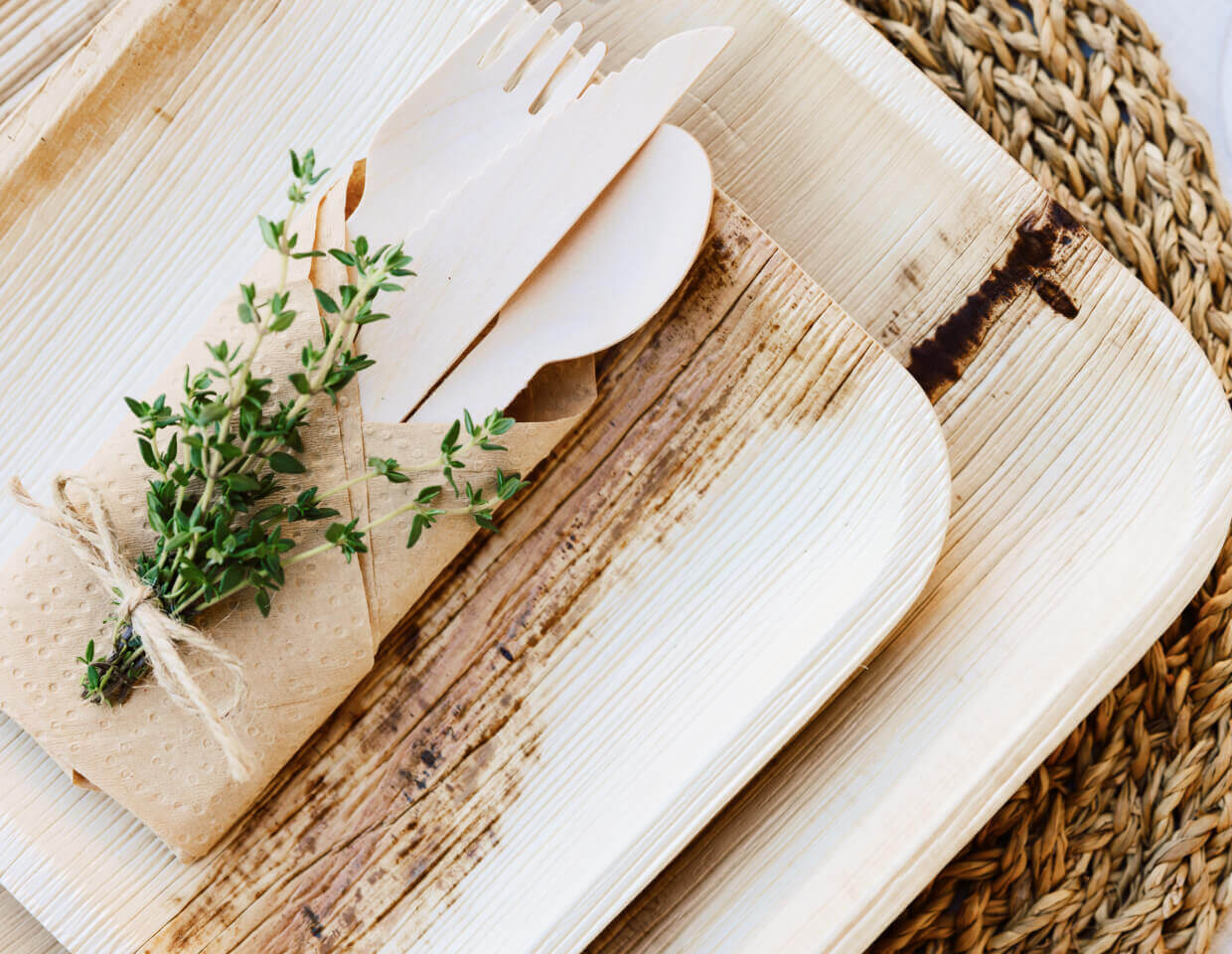 biodegradable ChicLeaf Palm Leaf Party Tablewear Set with palm leaf knives, spoons and forks a small plant and white ceramic items on a woven surface