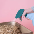 Person using a teal spray bottle to sprinkle cat litter into a brown litter box on a pink background